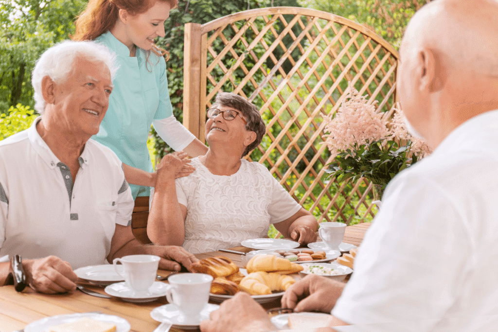 A tender caregiver assisting an elderly woman during a snack time outside on the patio of a happy care home for seniors.